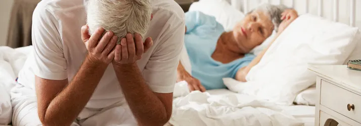 A man sitting at the edge of his bed, holding his forehead.